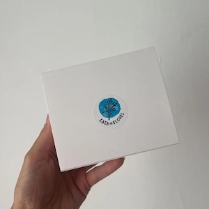 Puede incluir: Una caja cuadrada blanca con una pegatina circular en el centro. La pegatina muestra un fondo azul estilo acuarela con un dibujo lineal negro de un &aacute;rbol y el texto CASA AND FLORES impreso en letras negras a lo largo del borde inferior.