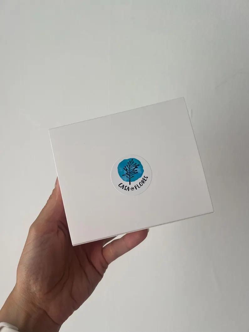 Puede incluir: Una caja cuadrada blanca con una pegatina circular en el centro. La pegatina muestra un fondo azul estilo acuarela con un dibujo lineal negro de un &aacute;rbol y el texto CASA AND FLORES impreso en letras negras a lo largo del borde inferior.
