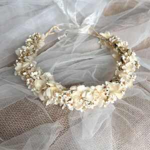May include: A circular floral crown featuring cream-colored dried hydrangea petals and small white flowers with yellow centers. The arrangement is accented with tiny metallic gold beads and is secured with a thin, translucent white ribbon tie.
