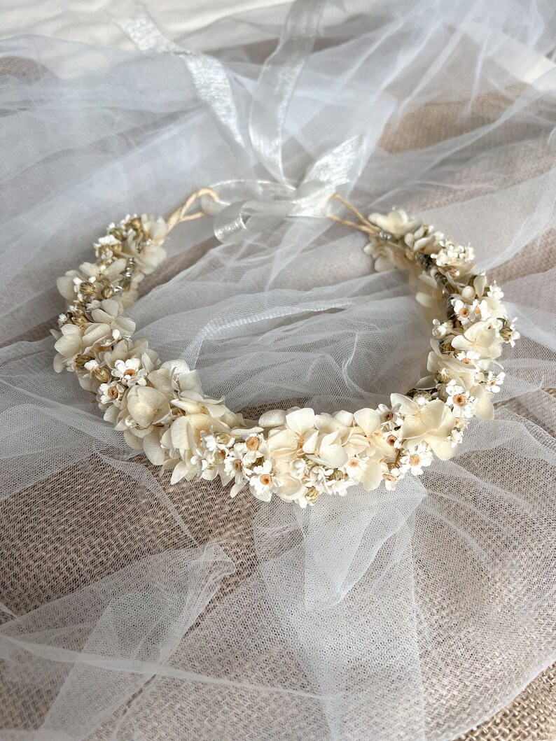 May include: A circular floral crown featuring cream-colored dried hydrangea petals and small white flowers with yellow centers. The arrangement is accented with tiny metallic gold beads and is secured with a thin, translucent white ribbon tie.