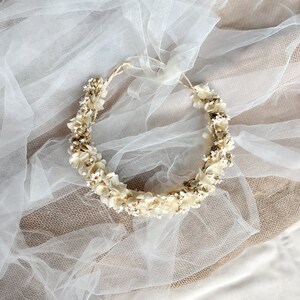 May include: A circular floral crown featuring small, cream-colored dried flowers and delicate white blossoms arranged on a thin, flexible base. The accessory includes a long, sheer white organza ribbon tied at the top for adjustable fastening.
