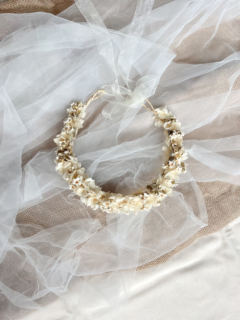 May include: A circular floral crown featuring small, cream-colored dried flowers and delicate white blossoms arranged on a thin, flexible base. The accessory includes a long, sheer white organza ribbon tied at the top for adjustable fastening.