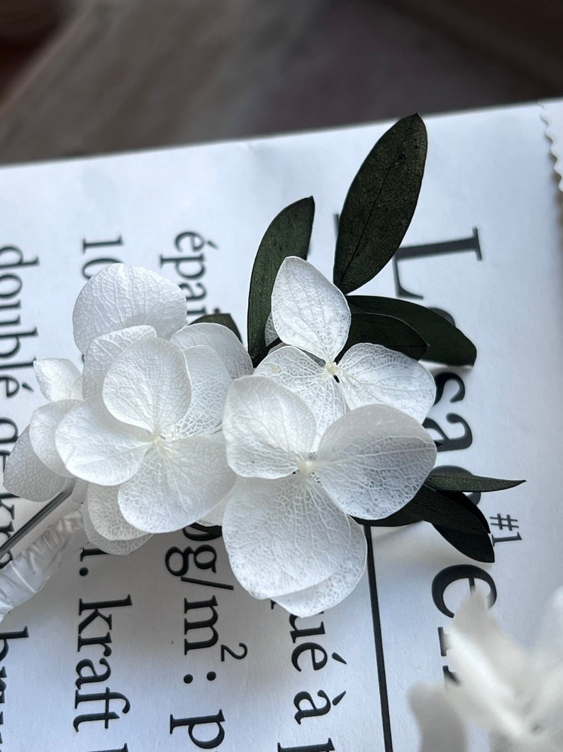 Puede incluir: Un delicado prendedor con p&eacute;talos de hortensia blancos con venas visibles y hojas de color verde oscuro. El arreglo floral est&aacute; sujeto a un tallo de pl&aacute;stico transparente y descansa sobre una superficie de papel blanco con texto impreso en negro.