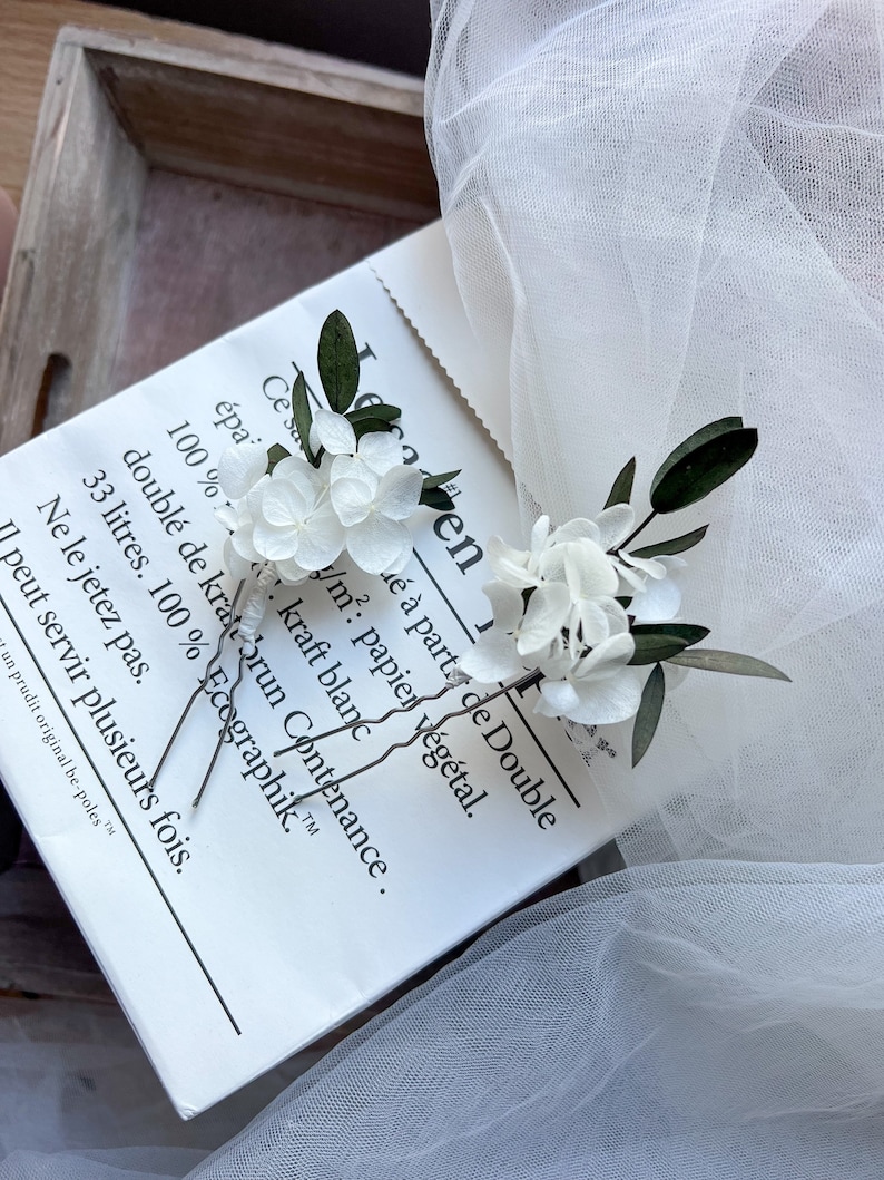 Puede incluir: Dos horquillas para el cabello con racimos de peque&ntilde;as flores de hortensia blancas y hojas de color verde oscuro. Cada horquilla tiene una base met&aacute;lica fina en forma de U, envuelta en cinta floral blanca para una sujeci&oacute;n segura y delicada.