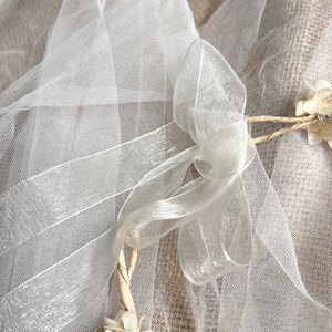 May include: A white sheer tulle veil featuring a delicate organza ribbon tied into a bow. The veil is accented with small, dried cream-colored flowers attached to a thin, twisted natural fiber vine frame, creating a soft and layered appearance.