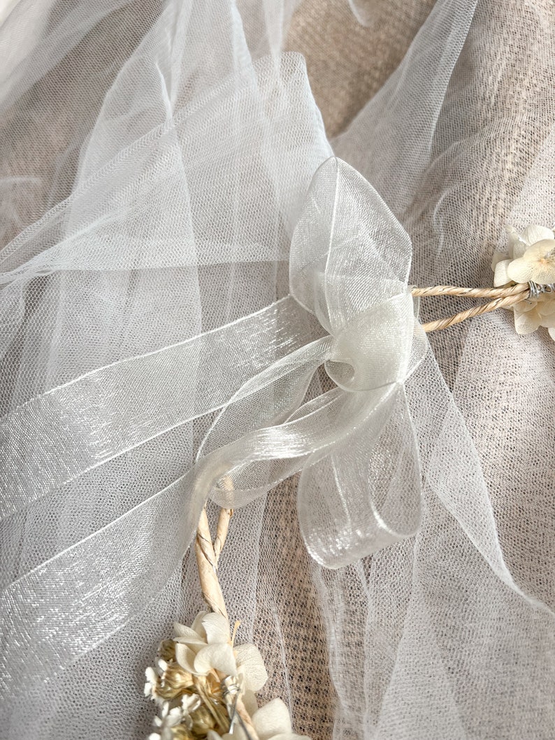 May include: A white sheer tulle veil featuring a delicate organza ribbon tied into a bow. The veil is accented with small, dried cream-colored flowers attached to a thin, twisted natural fiber vine frame, creating a soft and layered appearance.