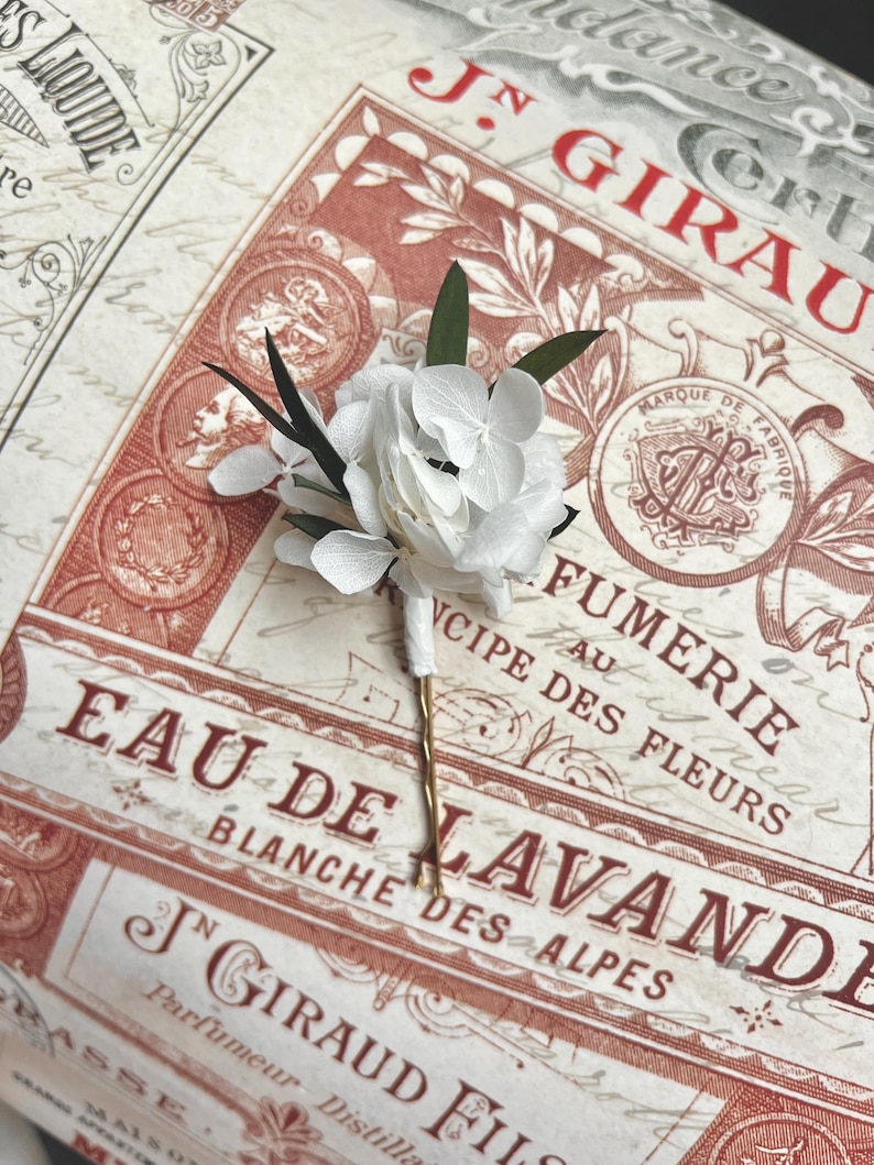 Puede incluir: Una horquilla para el pelo con un ramillete de p&eacute;talos de hortensia blancos y peque&ntilde;as hojas de color verde oscuro. El tallo del arreglo floral est&aacute; envuelto en cinta blanca y unido a una fina horquilla de metal dorado.