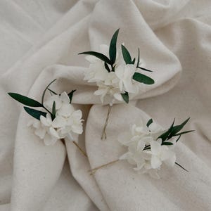 Puede incluir: Tres horquillas para el pelo con racimos de peque&ntilde;as flores blancas acentuadas con hojas finas de color verde oscuro. Cada arreglo floral est&aacute; unido a un alfiler de metal fino, dise&ntilde;ado para insertarse en el cabello como adorno floral.