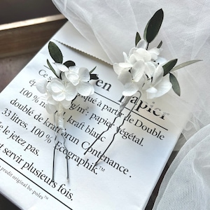 Puede incluir: Dos horquillas para el cabello con p&eacute;talos de hortensia blancos y peque&ntilde;as hojas de color verde oscuro. Los tallos de las horquillas est&aacute;n envueltos en hilo blanco y las puntas de metal son plateadas. El arreglo floral es delicado y natural.