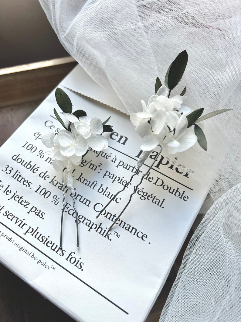 Puede incluir: Dos horquillas para el cabello con p&eacute;talos de hortensia blancos y peque&ntilde;as hojas de color verde oscuro. Los tallos de las horquillas est&aacute;n envueltos en hilo blanco y las puntas de metal son plateadas. El arreglo floral es delicado y natural.