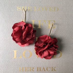 May include: A pair of earrings featuring two burgundy red paper flower designs. The flowers are layered and have a delicate, ruffled texture. The earrings are suspended from gold-toned hooks.