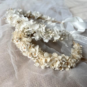 May include: Two floral crowns constructed from preserved white and cream-colored hydrangea petals and small filler flowers. Each circular headpiece features a thin, flexible base and is finished with long, smooth white satin ribbons for tying.
