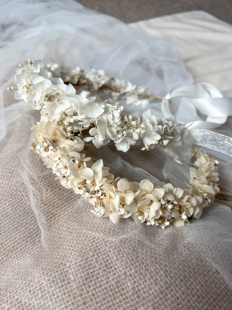 May include: Two floral crowns constructed from preserved white and cream-colored hydrangea petals and small filler flowers. Each circular headpiece features a thin, flexible base and is finished with long, smooth white satin ribbons for tying.