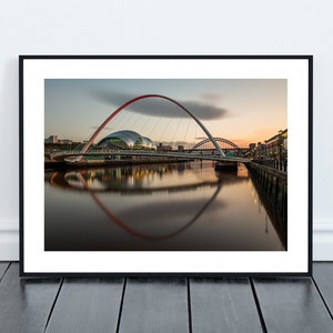 May include: A red and white arch bridge over a river with a reflection of the bridge in the water. The bridge is lit up at night and there are buildings in the background.
