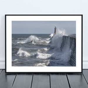 May include: A black and white photograph of a lighthouse on a rocky pier with waves crashing against it. The lighthouse is tall and slender, with a white tower and a black roof. The waves are large and powerful, and they are crashing against the pier with great force. The photograph is taken from a low angle, so the waves appear to be towering over the lighthouse.