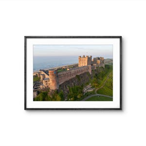 May include: Framed print of a historic castle perched on a cliff overlooking the ocean. The castle is made of stone and has multiple towers and walls. The sky is a soft blue, and the water is calm.