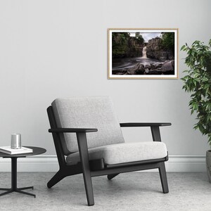 May include: A black armchair with a gray fabric seat and back, positioned in front of a white wall with a framed photograph of a waterfall in a forest setting.