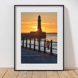 May include: A black and white photograph of a lighthouse silhouetted against a sunrise. The lighthouse is on a pier with a railing and a life ring hanging from it. The water is calm and the sky is a vibrant orange and yellow.