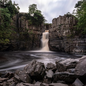 May include: A waterfall cascades over a rocky cliff face, surrounded by lush green trees. The water flows into a pool below, creating a swirling pattern.