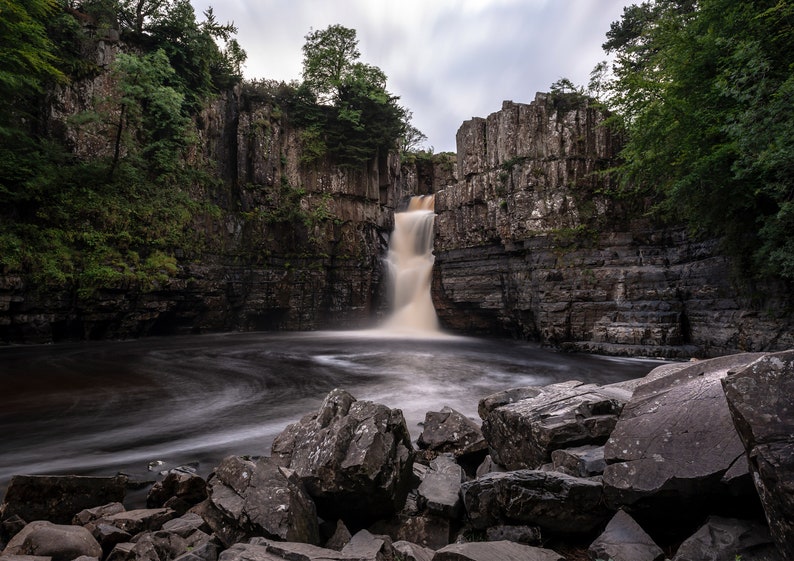May include: A waterfall cascades over a rocky cliff face, surrounded by lush green trees. The water flows into a pool below, creating a swirling pattern.