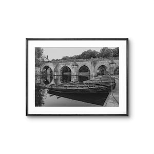 May include: Black and white framed print of a stone bridge with multiple arches reflected in the water. Several wooden boats are docked in the foreground. The image is a landscape photograph.