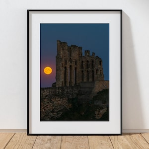 May include: Framed print of a castle ruin silhouetted against a twilight sky. A large, orange full moon is positioned near the castle. The print is in a black frame and rests on a wooden surface.
