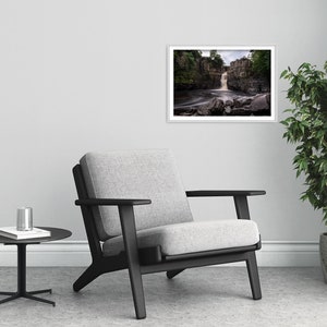 May include: A framed photograph of a waterfall cascading over rocks in a forest setting. The water is flowing over the rocks and creating a mist. The photograph is in a white frame and is hanging on a white wall.