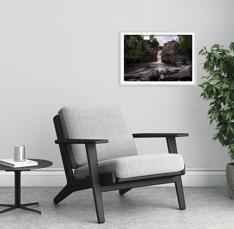 May include: A framed photograph of a waterfall cascading over rocks in a forest setting. The water is flowing over the rocks and creating a mist. The photograph is in a white frame and is hanging on a white wall.