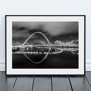 May include: Monochrome framed print of a bridge at night, featuring a prominent arch reflected in the water. The image captures the illuminated bridge against a dark, cloudy sky, creating a striking visual.