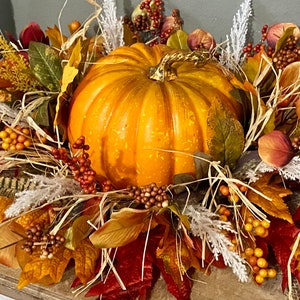 May include: A vibrant orange pumpkin centerpiece surrounded by fall foliage. The arrangement includes colorful leaves in shades of red, yellow, and green, along with berries and decorative accents. This autumn-themed decor is perfect for seasonal displays.