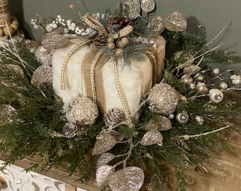 May include: A decorative holiday centerpiece featuring a plush, cream-colored square shape wrapped with burlap ribbon. The arrangement includes pinecones, silver glittered ornaments, and evergreen foliage, creating a festive and elegant display.