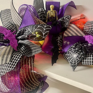 May include: A Halloween-themed decorative wreath with purple, black, and white ribbons. The wreath features spiderwebs, bats, and a small skeleton. The design includes lights for added effect, creating a festive and spooky atmosphere.