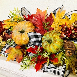 May include: A fall-themed decorative arrangement featuring pumpkins, colorful leaves, berries, pinecones, and black and white checkered ribbon. The pumpkins are orange and green, and the leaves are red, yellow, and brown.