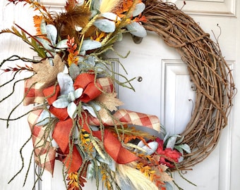 May include: A grapevine wreath adorned with fall-themed faux foliage. The wreath features orange, brown, and green leaves, wheat, and a large bow with a plaid pattern. The wreath is a decorative piece for the front door.