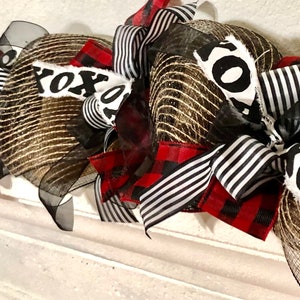 May include: A gold and black mesh wreath with red and black buffalo plaid ribbon and black and white striped ribbon. The wreath has a white fabric sign that says "XOXO".
