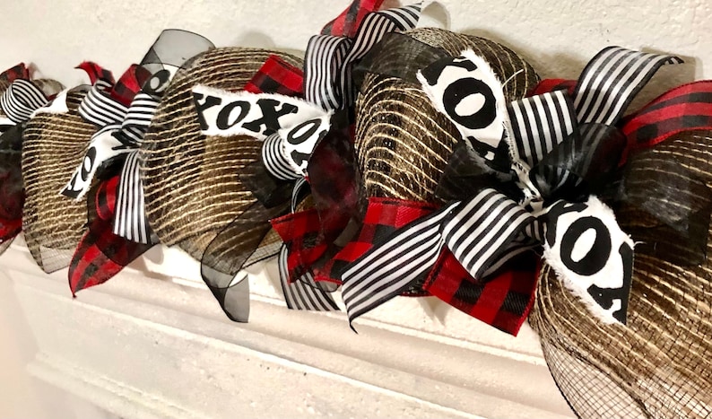 May include: A gold and black mesh wreath with red and black buffalo plaid ribbon and black and white striped ribbon. The wreath has a white fabric sign that says "XOXO".