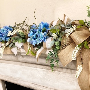 May include: A blue and white floral garland with burlap bows and greenery. The garland is displayed on a white fireplace mantel.