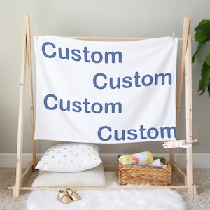 May include: A white blanket with the word "Custom" written four times in blue lettering, hanging on a wooden stand. The blanket is surrounded by pillows and a basket with a colorful blanket inside.