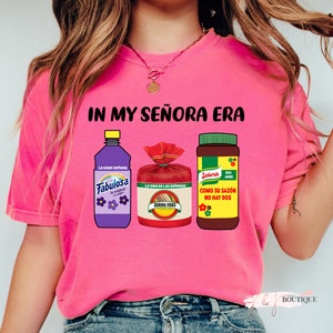 May include: Pink t-shirt with the text "IN MY SE&Ntilde;ORA ERA" and images of three products: a purple bottle of Fabulosa cleaner, a red bag of Se&ntilde;ora Vibes seasoning, and a brown bottle of Se&ntilde;ora seasoning.
