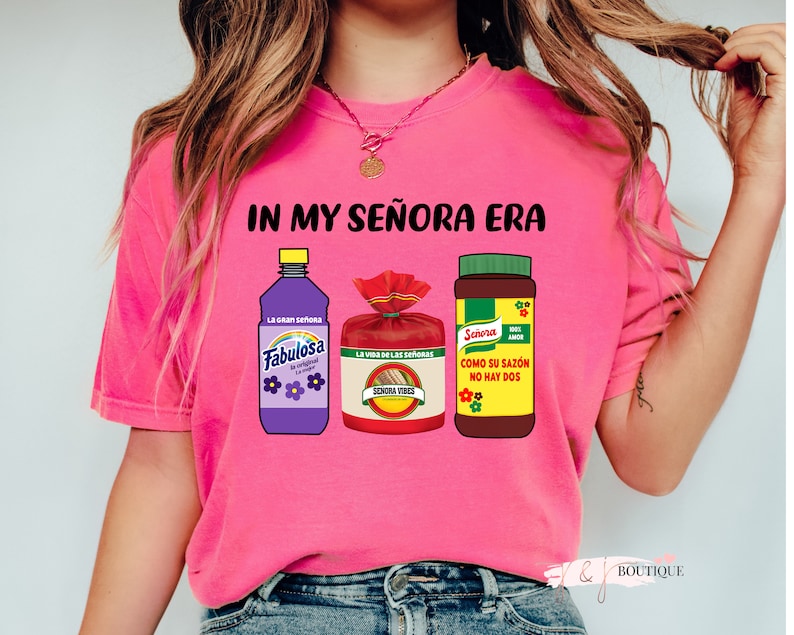 May include: Pink t-shirt with the text "IN MY SE&Ntilde;ORA ERA" and images of three products: a purple bottle of Fabulosa cleaner, a red bag of Se&ntilde;ora Vibes seasoning, and a brown bottle of Se&ntilde;ora seasoning.