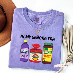 May include: Purple t-shirt with the text "IN MY SE&Ntilde;ORA ERA" and illustrations of three products: Fabulosa, La Viga de las Se&ntilde;oras, and Saz&oacute;n.