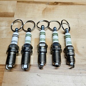 May include: Five spark plug keychains with silver metal rings. The spark plugs are silver with white and green accents and the word "BOSCH" printed on them. The keychains are arranged on a light brown wooden surface.