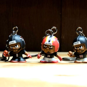 May include: Three Atlanta Falcons football player keychains. Each keychain features a miniature player figurine in a black and red uniform, wearing a helmet with the Falcons logo. The figurines are on a wooden surface.