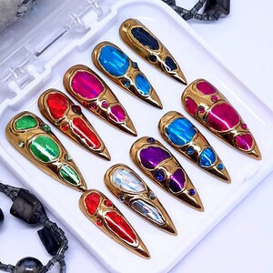 May include: A collection of ten gold-toned nail art pieces, each featuring a large, colorful gemstone. The gemstones are in various colors, including blue, pink, red, green, and purple. The nails are arranged in a white tray.