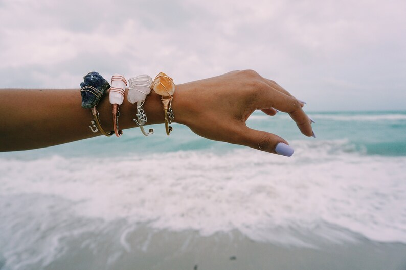 May include: A person's wrist wearing five bracelets with different colored stones. The bracelets are made of wire wrapped around the stones. The person's hand is reaching out towards the ocean.