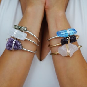 May include: Three silver and gold wire wrapped bracelets with different crystals. The top bracelet features a light blue crystal, the middle bracelet features a black crystal, and the bottom bracelet features a pink crystal.