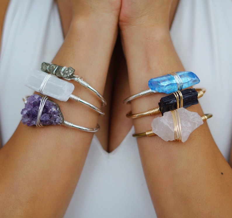 May include: Three silver and gold wire wrapped bracelets with different crystals. The top bracelet features a light blue crystal, the middle bracelet features a black crystal, and the bottom bracelet features a pink crystal.