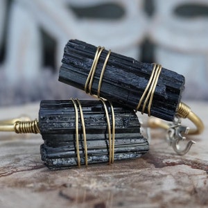 May include: Two black tourmaline crystal bracelets wrapped in gold wire. The bracelets are on a wooden surface. One bracelet has an Om symbol charm.
