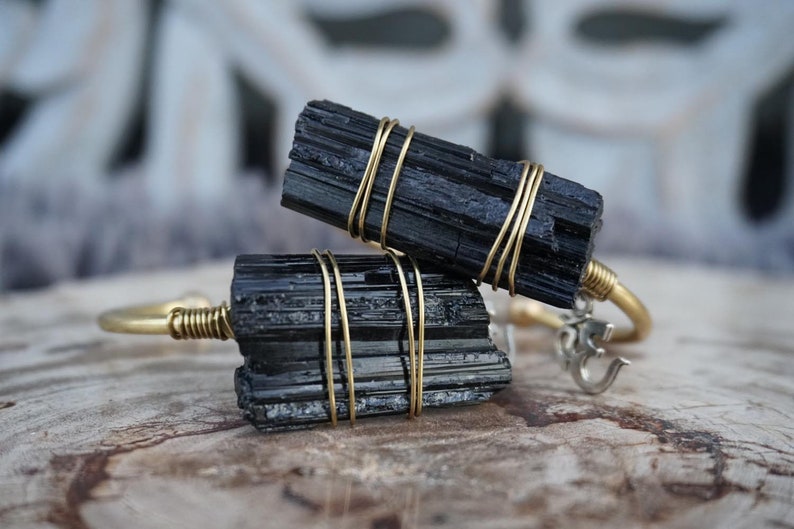 May include: Two black tourmaline crystal bracelets wrapped in gold wire. The bracelets are on a wooden surface. One bracelet has an Om symbol charm.