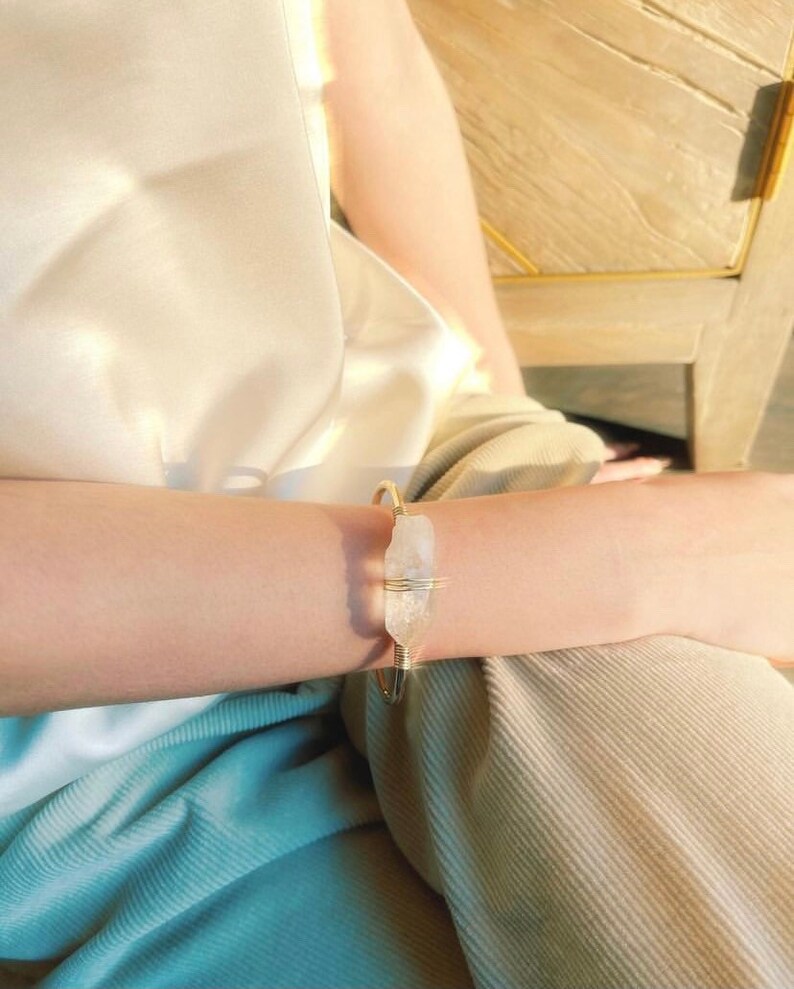 May include: A gold wire bracelet with a clear quartz crystal wrapped in the wire. The bracelet is on a person's wrist.